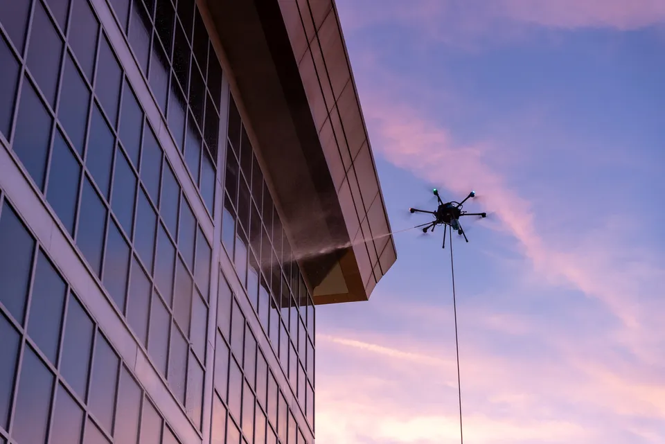 Lucid Bots Sherpa cleaning drone spraying soft-wash solution across a glass building corner at dusk